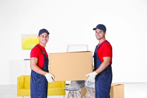 Two male movers dressed in red shirts, blue overalls, and white gloves are standing indoors against a plain white wall, holding a large cardboard box together. One is positioned on the left, and the other on the right, both smiling and looking toward the camera. Behind them, there is a yellow upholstered sofa with a modern design, placed on a light-colored wooden floor. On the wall behind, a framed piece of artwork featuring yellow and green abstract elements is partially visible. To the right of the movers, there is a small wooden side table with a grey cloth draped over it, supporting the bottom of the box they are carrying. Additionally, a closed cardboard box is placed on the floor near the sofa, indicating a typical house removal environment. The lighting appears bright and even, emphasizing the clean, uncluttered interior suitable for house removals and relocation services in the Dulwich area, providing a clear visual context of professional movers engaged in packing or loading household items.