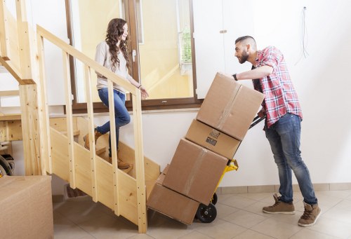 A man with a beard, wearing a red and white checkered shirt and jeans, is loading three cardboard boxes onto a black hand truck inside a residential hallway, assisting a woman with long, curly brown hair, dressed in a white top and blue jeans, who is standing at the bottom of a wooden staircase with natural finish. The boxes, which are sealed with packing tape, are stacked on the hand truck, showing a logo on the top box. The hallway has white walls, a large window allowing natural light, and beige tiled flooring. A second cardboard box rests on the floor in the foreground. The environment appears to be part of a house in Dulwich or nearby, with a focus on home relocation and removals services provided by Dulwich Man with Van, emphasizing a careful and organized moving process.