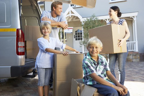 A family engaged in a house removal in a residential driveway, with three members actively involved in packing and loading boxes into a large, grey removals van. An adult woman, dressed in a striped shirt and jeans, holds a medium-sized cardboard box, while a man leans on the open rear door of the van, watching and smiling. Two children, a girl in a light blue shirt and denim shorts and a boy in a plaid shirt and jeans, are positioned near stacks of cardboard boxes, with the girl resting her hand on a box and the boy seated on a large box, surrounded by additional packing cartons. The scene takes place outside a detached house with white siding and dark window frames, indicating a suburban environment, possibly within Dulwich, South London. The lighting appears natural, likely early afternoon, highlighting the family's cheerful mood during the moving process, which involves household items such as cardboard boxes, packing materials, and personal belongings. Dulwich Man with Van provides professional removals services, facilitating efficient house relocations within the local area, as illustrated in this residential scene.